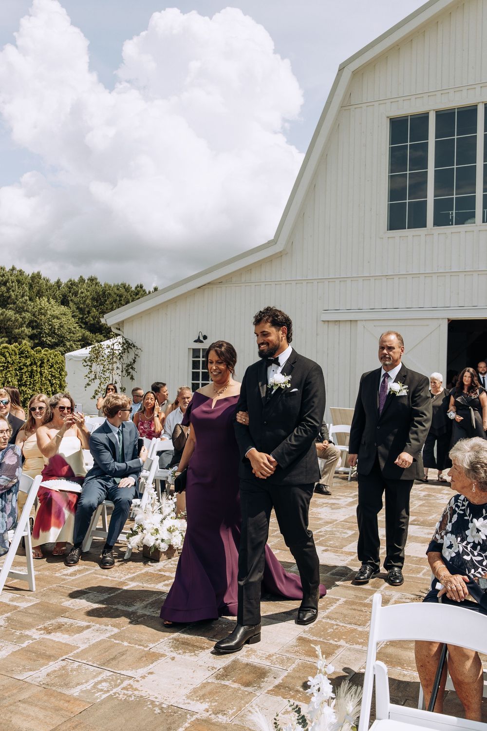 Groom escorts mother down outdoor wedding aisle at white barn venue on sunny day.