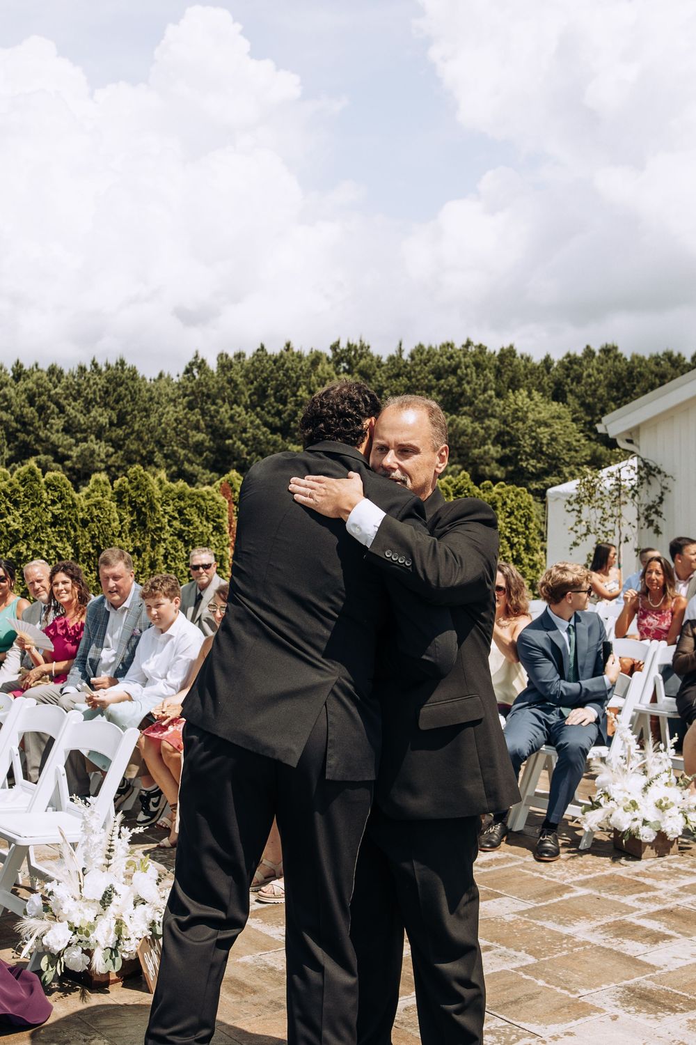 Two men in black suits share heartfelt embrace at outdoor wedding ceremony.