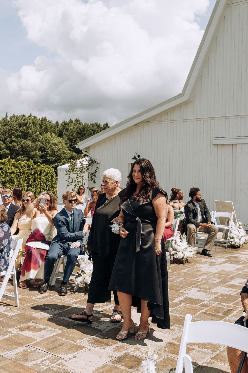Two women in black dresses walk down aisle at elegant outdoor barn wedding.