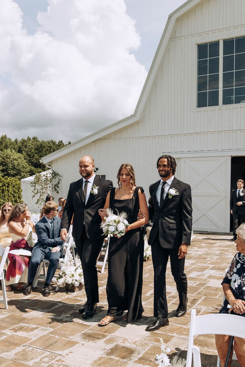 Bridesmaid in black gown escorted by two groomsmen down outdoor wedding aisle.