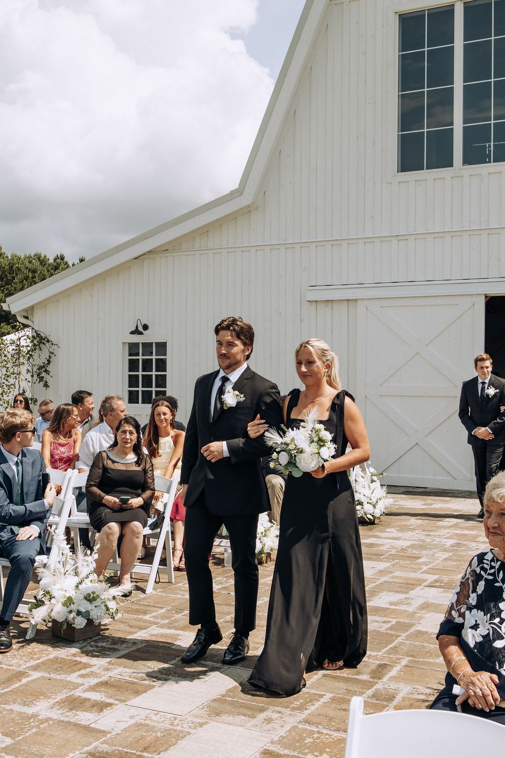 Groomsman in black suit escorts bridesmaid holding white bouquet down wedding aisle.