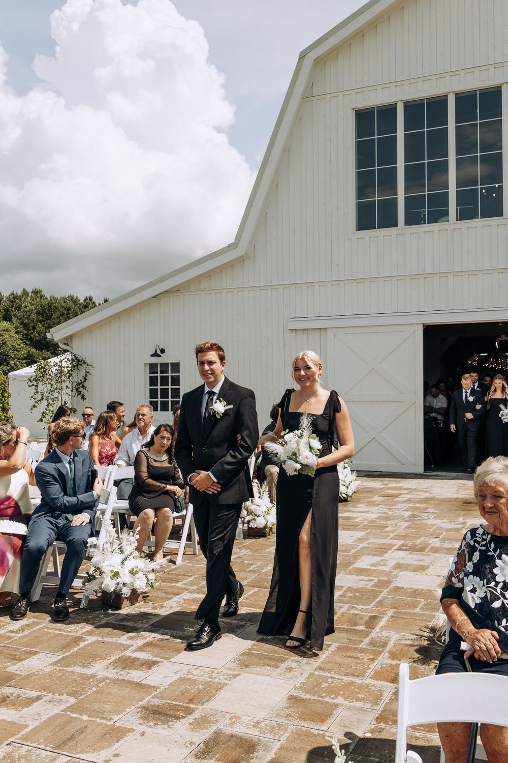 Groomsman escorts bridesmaid in black gown with white bouquet down outdoor aisle.