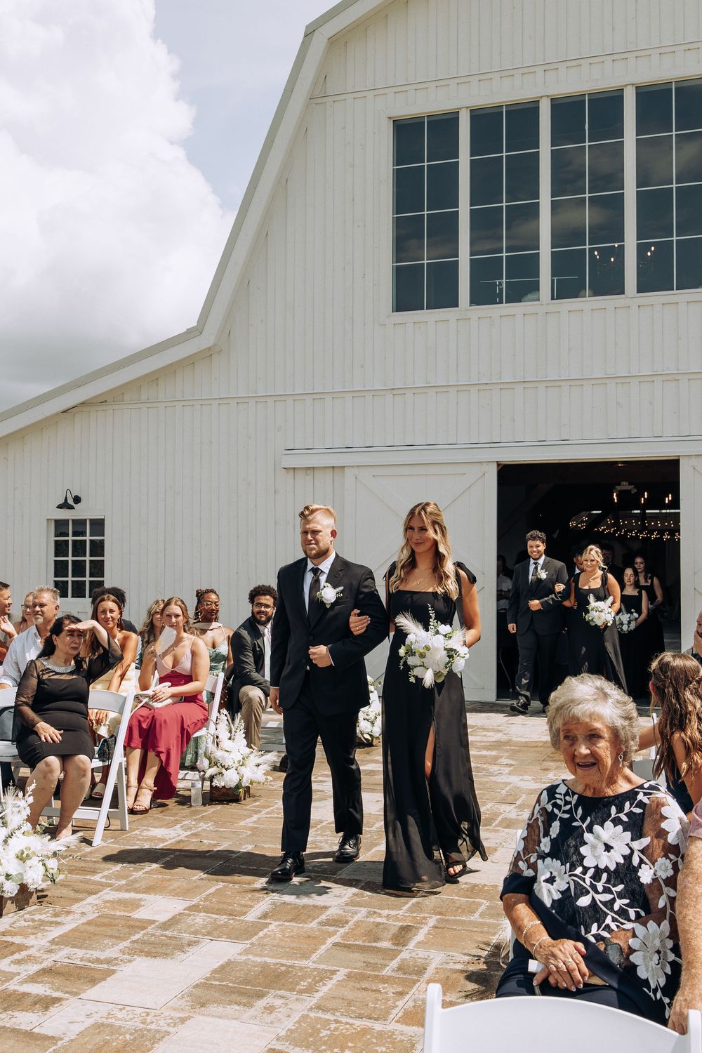 Groomsman escorts bridesmaid carrying white bouquet down sunny outdoor barn wedding aisle.