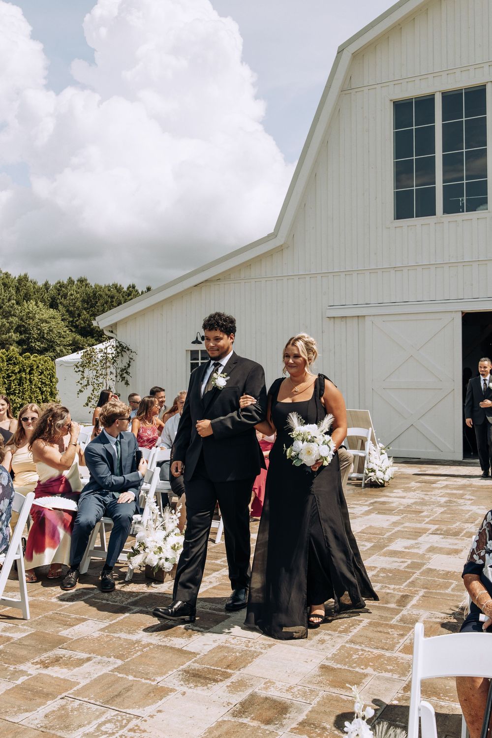 Groomsman escorts bridesmaid in black dress holding white bouquet down wedding aisle.
