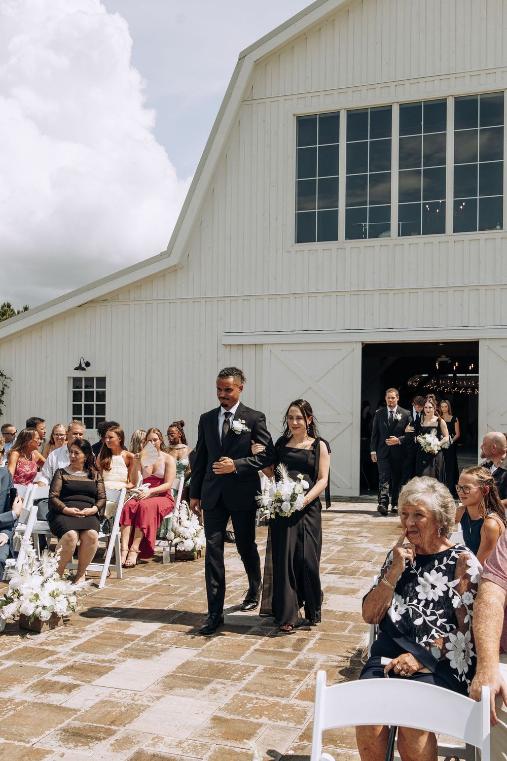 Groomsman escorts bridesmaid in black gown down outdoor barn wedding ceremony aisle.