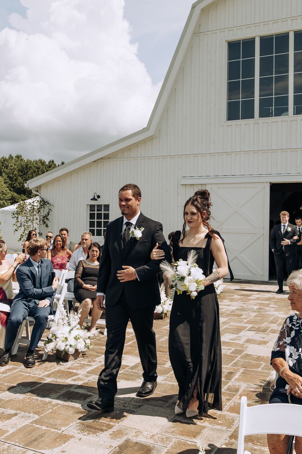 Groomsman escorts bridesmaid in black dress down outdoor wedding aisle at white barn venue.
