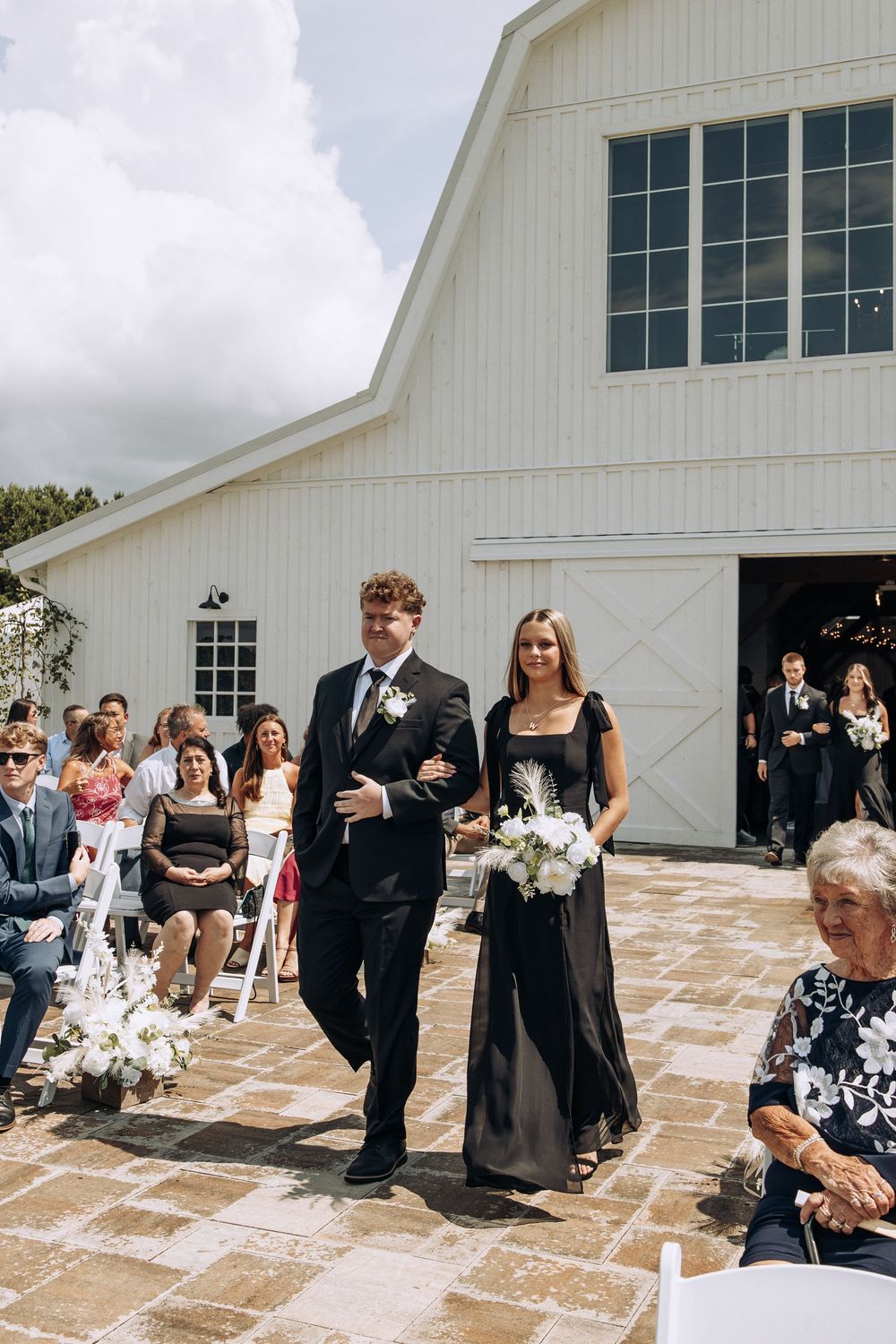 Groomsman with curly hair escorts bridesmaid in black gown down sunlit outdoor wedding aisle.
