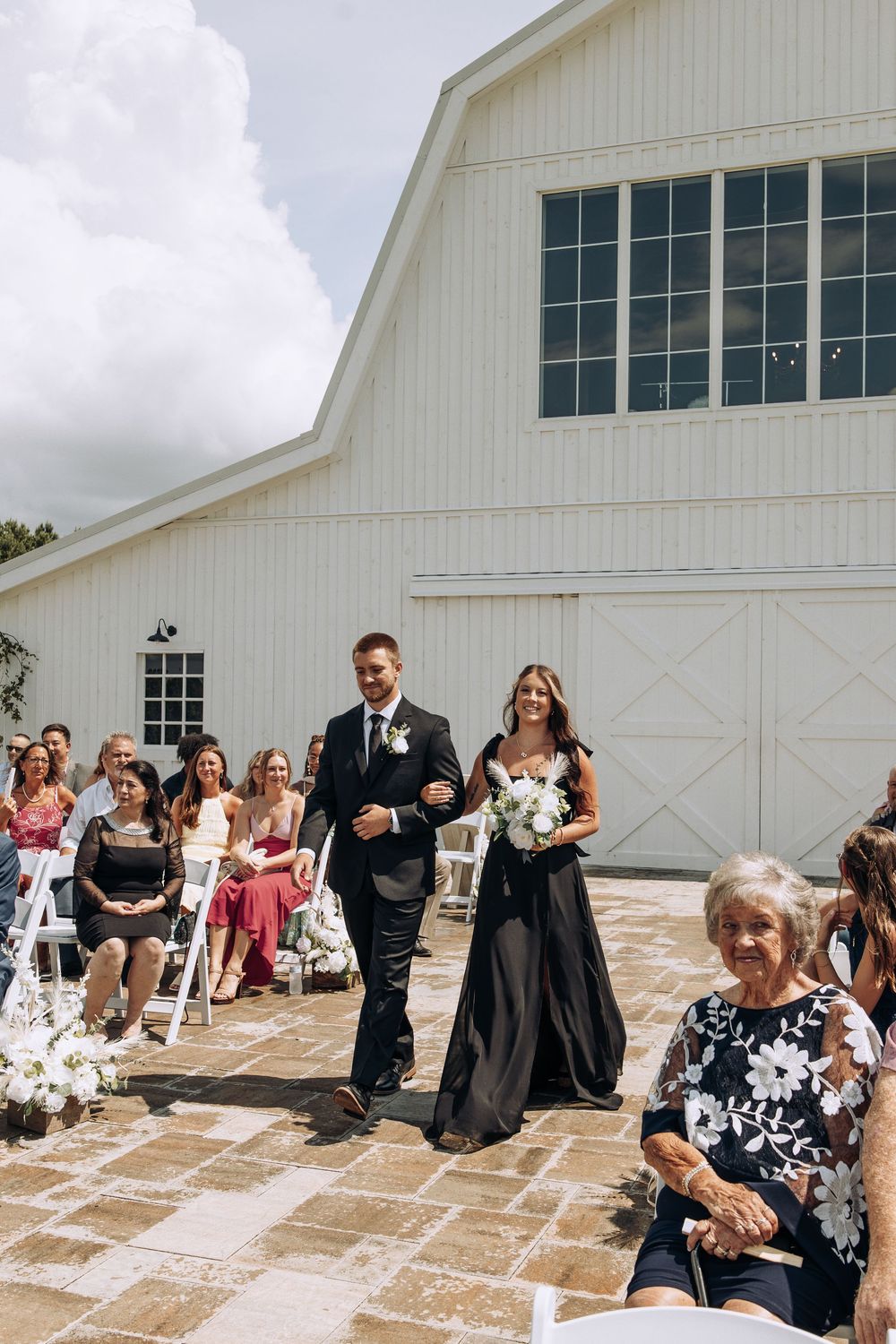 Smiling groomsman escorts bridesmaid in black dress down outdoor aisle at white barn wedding.