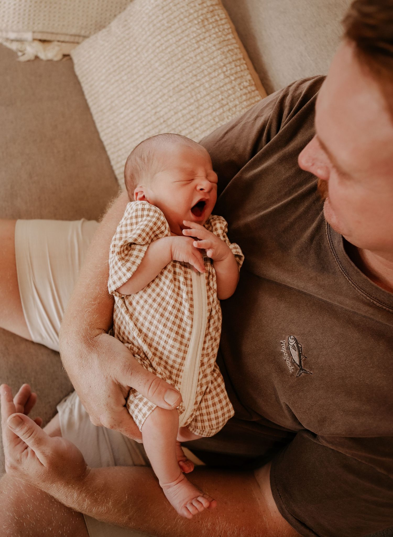A yawning newborn baby wearing a gingham outfit rests on someone's lap in a warm, natural light setting.