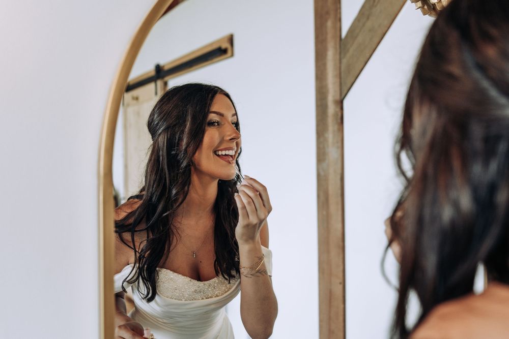 Smiling bride in white dress applying lipstick while looking into an arched wooden mirror.