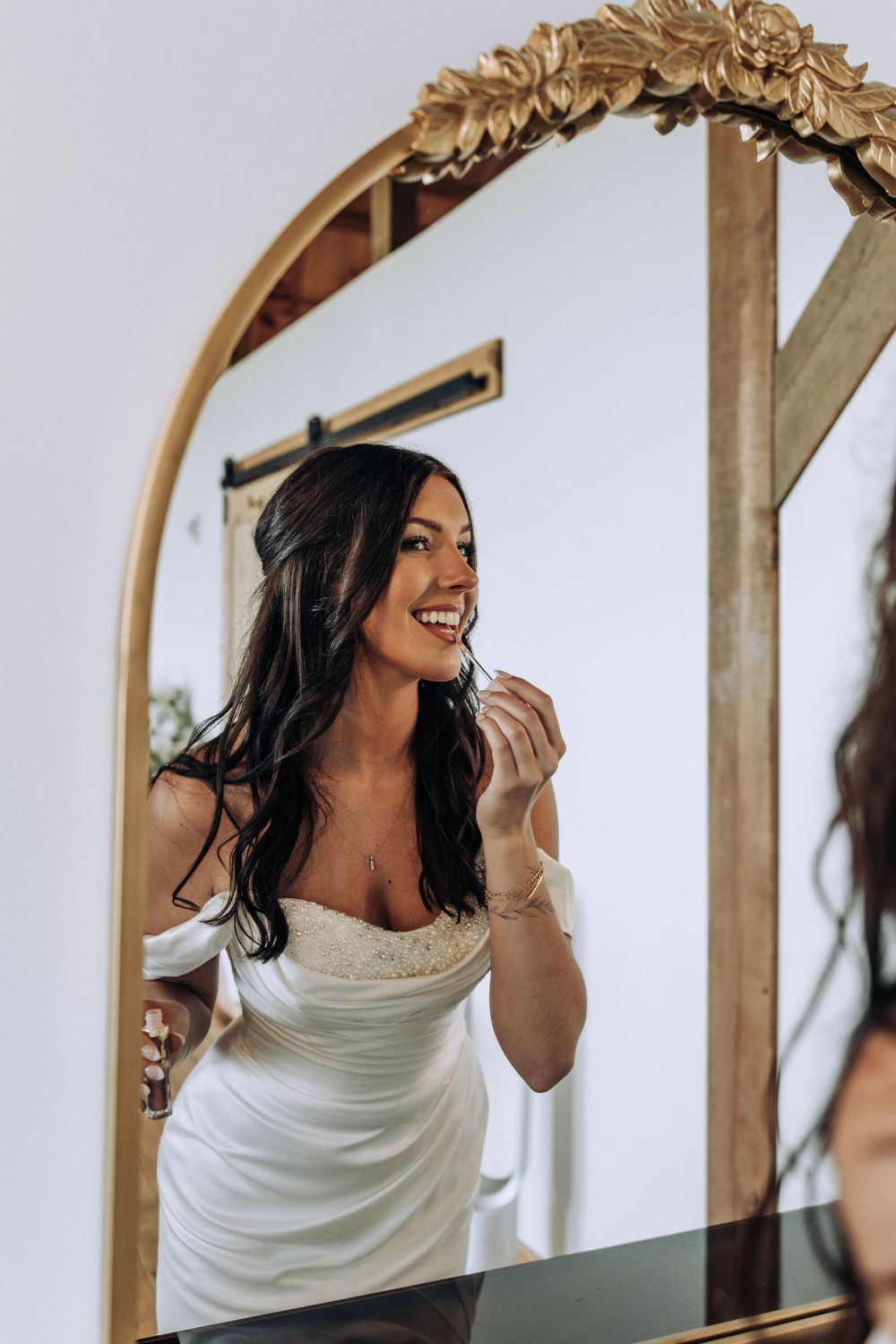 Beautiful bride in off-shoulder white dress smiling while applying lipstick in ornate gold mirror.
