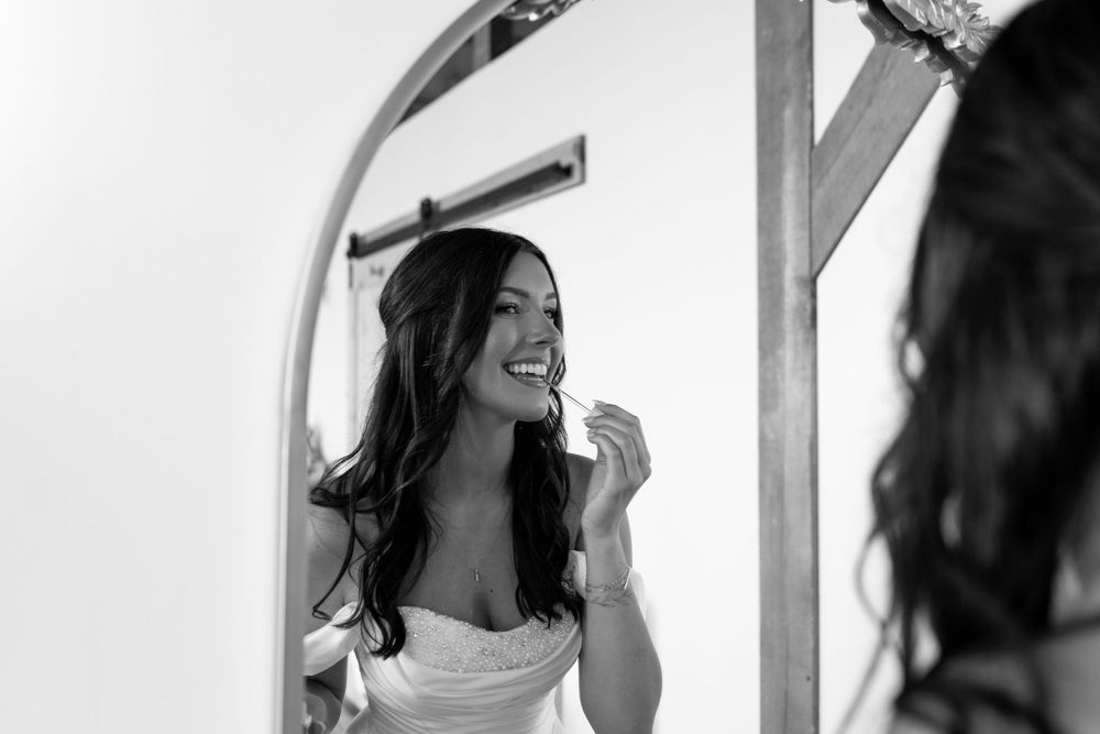 Black and white photo of a bride in white dress smiling while applying lipstick in a mirror.