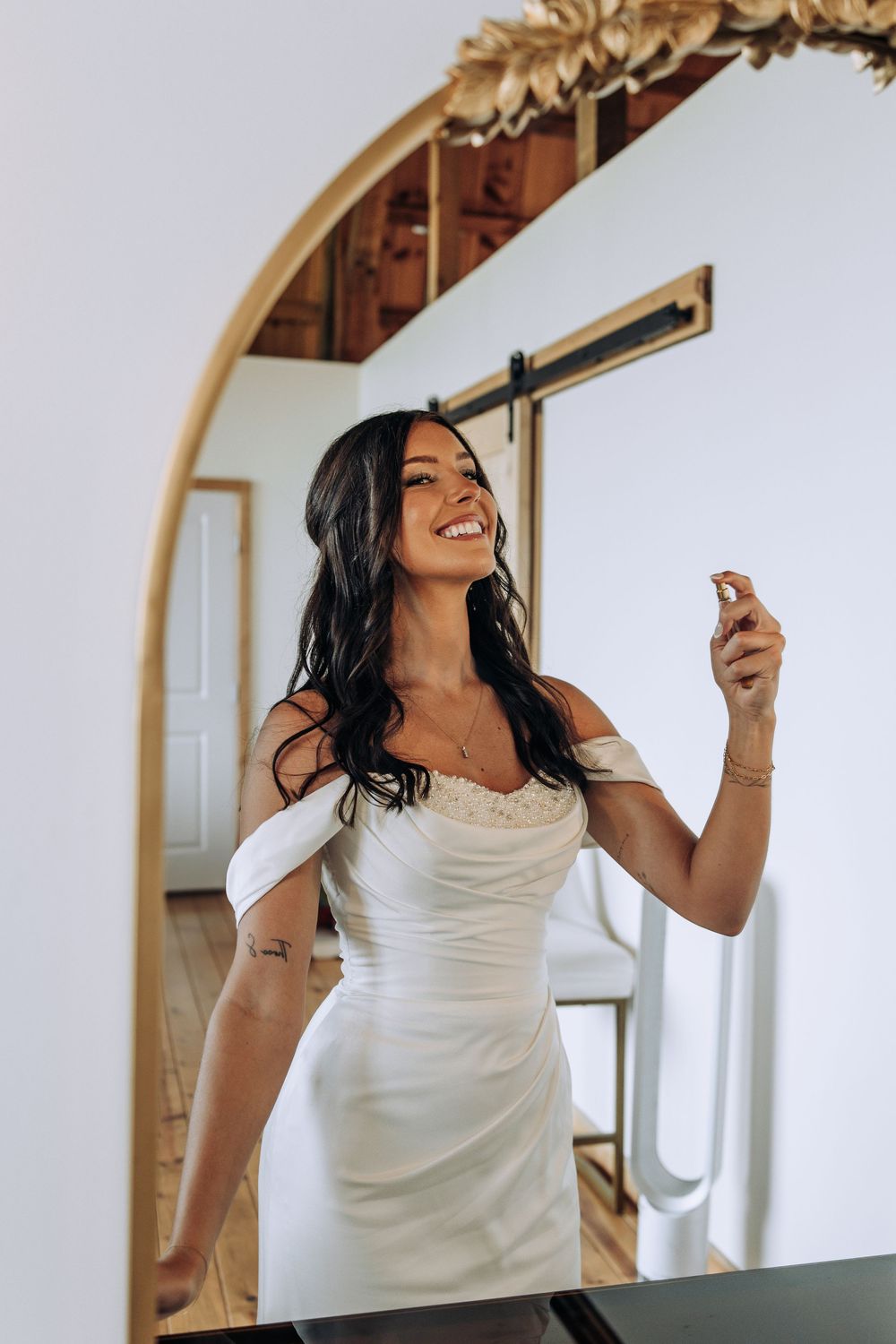 Smiling bride in white off-shoulder gown spraying perfume in a rustic venue with arched mirror.