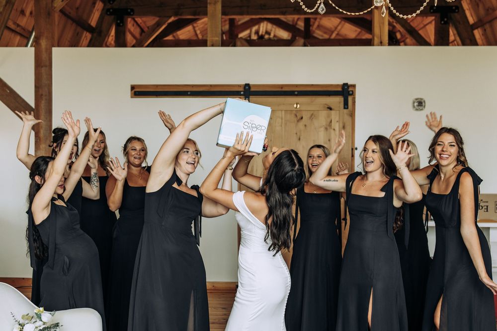 Excited bridesmaids in black dresses cheering with bride in white gown inside rustic barn venue.