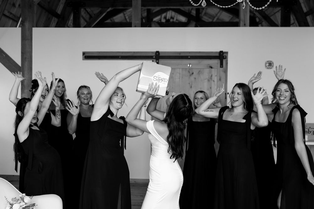 Black and white photo of bridesmaids in black dresses celebrating with bride in rustic barn setting.