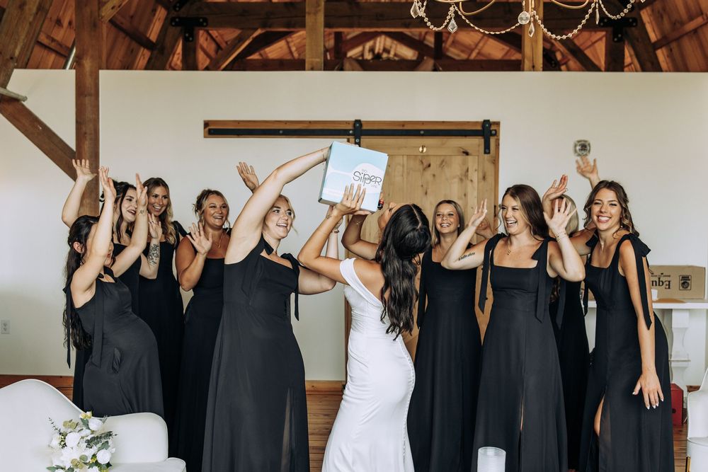 Bride in white gown holding book while bridesmaids in black dresses cheer in rustic barn venue.