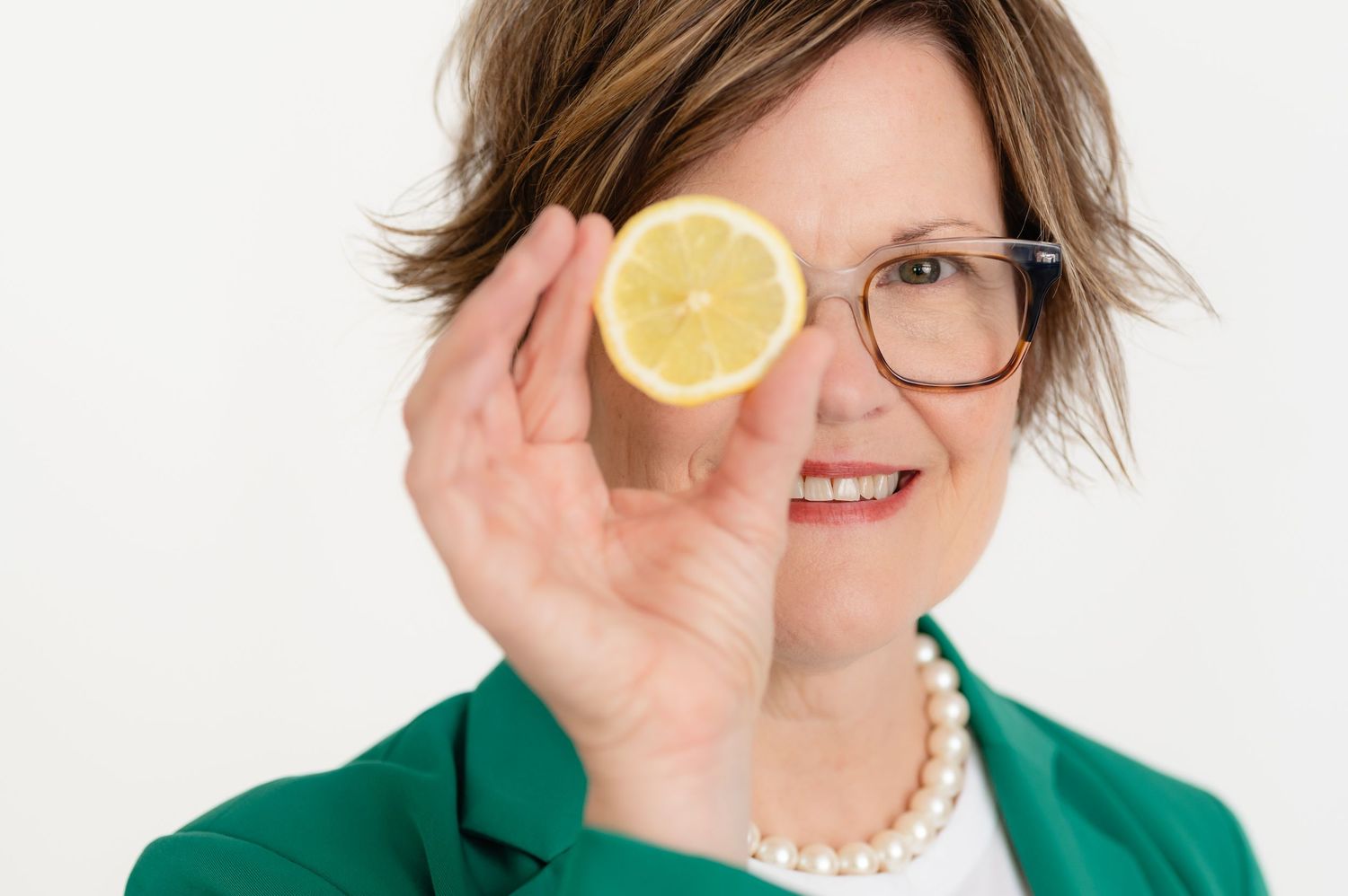Wellness coach Kathy Gregory in green blazer and glasses holds a halved lemon in front of their face against white background in Spring TX.