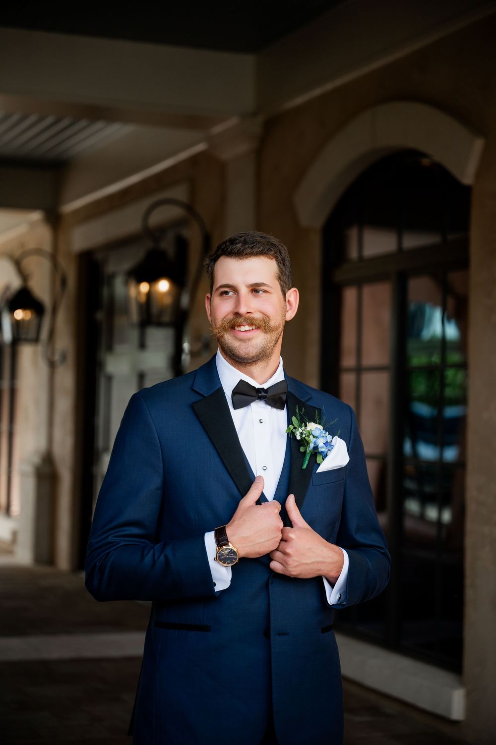 A groom in a navy blue tuxedo and black bow tie smiles while adjusting his jacket in an elegant venue.