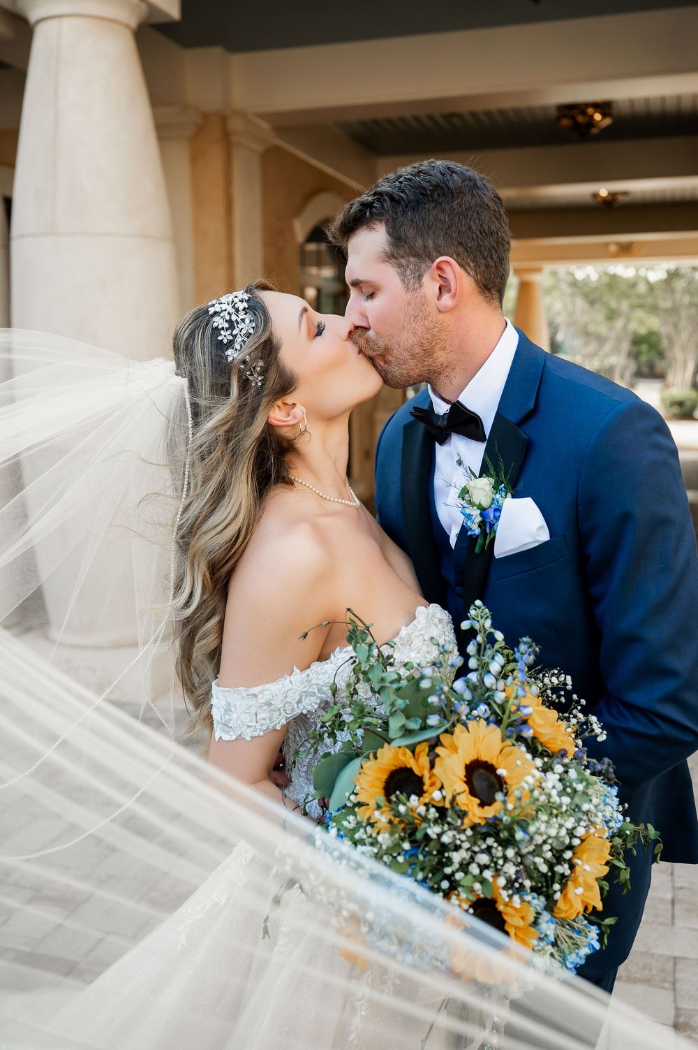 A wedding couple shares a romantic kiss while the bride holds a sunflower bouquet with baby's breath.
