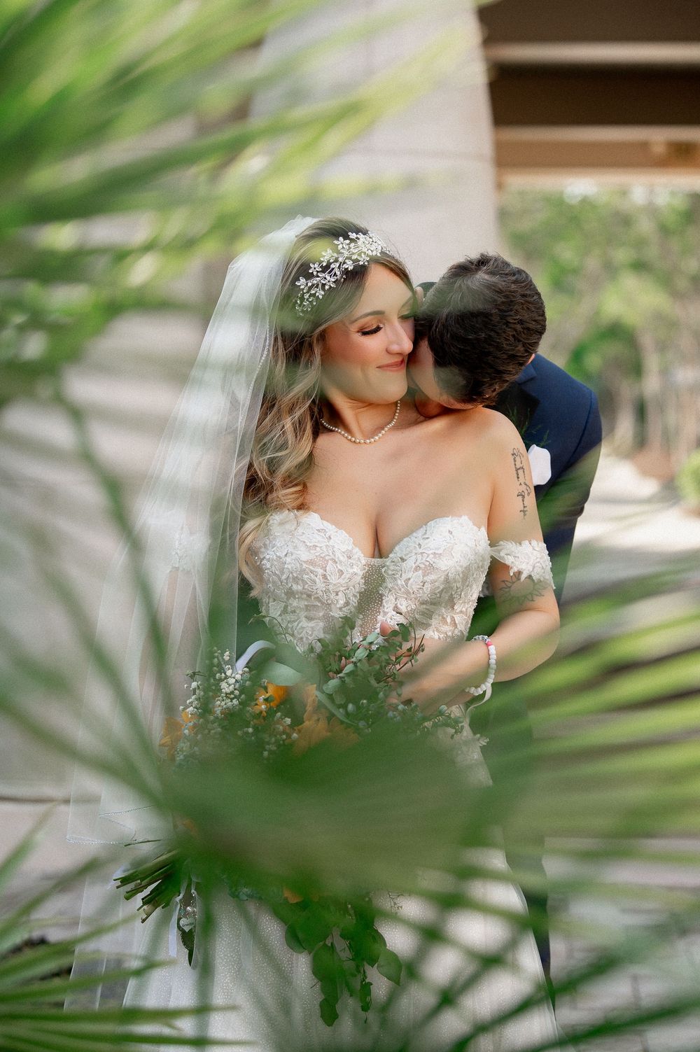 A romantic wedding portrait through palm leaves shows an intimate moment between newlyweds.