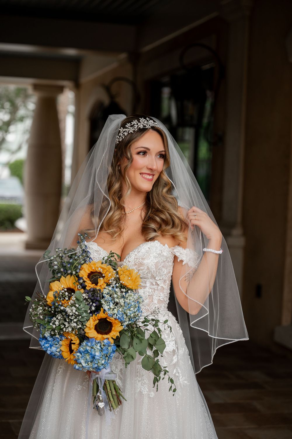 Bride in lace gown holds sunflower and hydrangea bouquet under columned archway.
