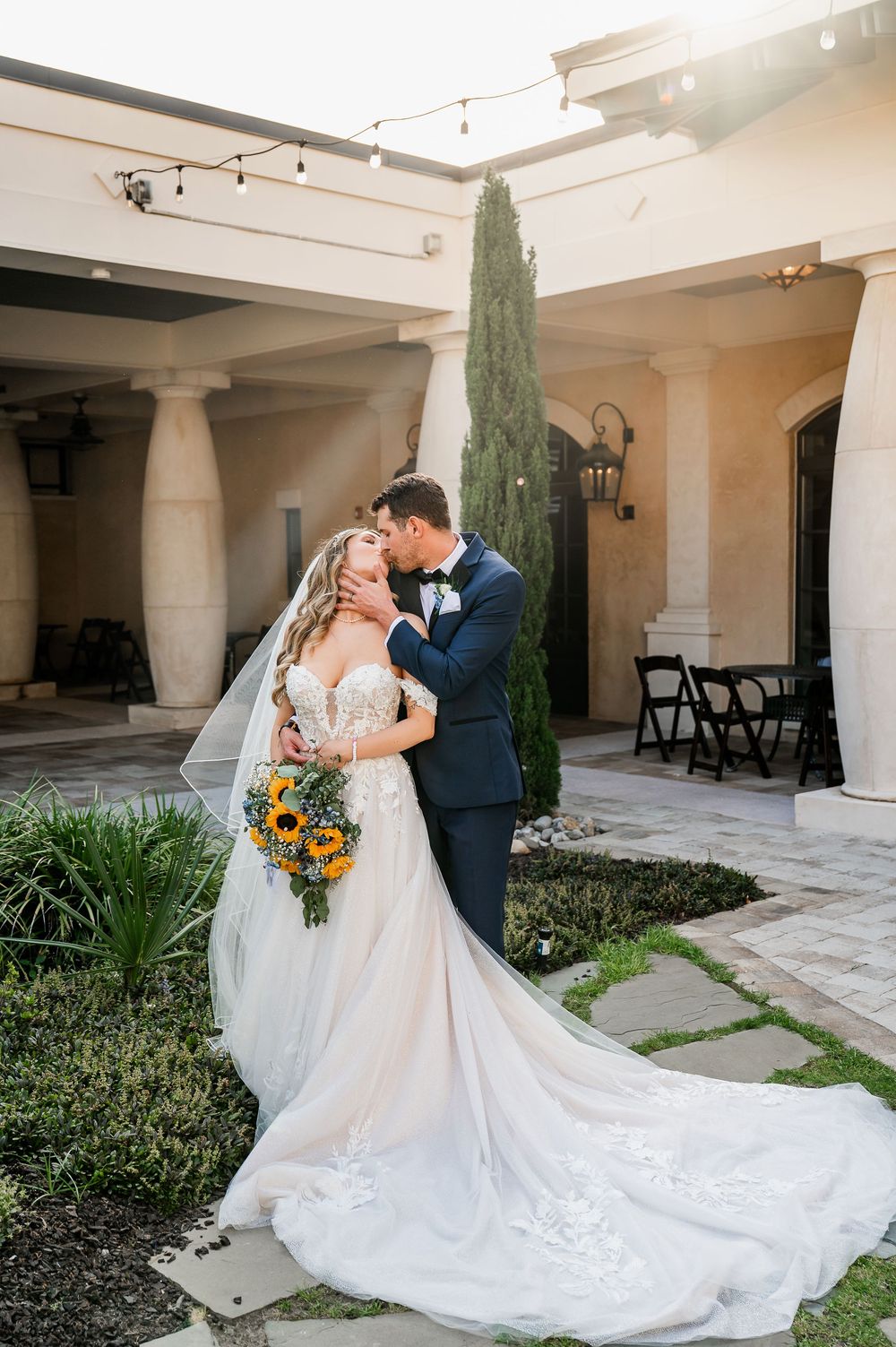 A romantic wedding portrait shows a couple embracing in front of a Mediterranean-style venue at sunset.