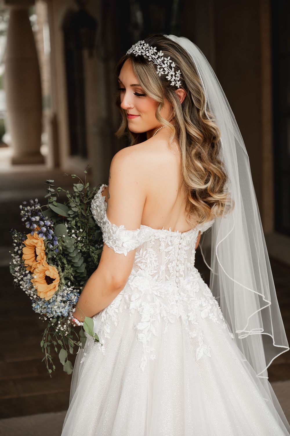 Back view of bride in off-shoulder wedding gown holding blue and yellow bouquet.