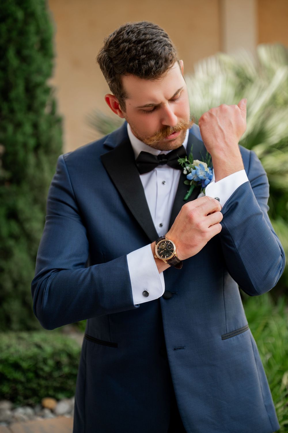 Groom in navy tuxedo adjusts cufflink while wearing luxury watch.