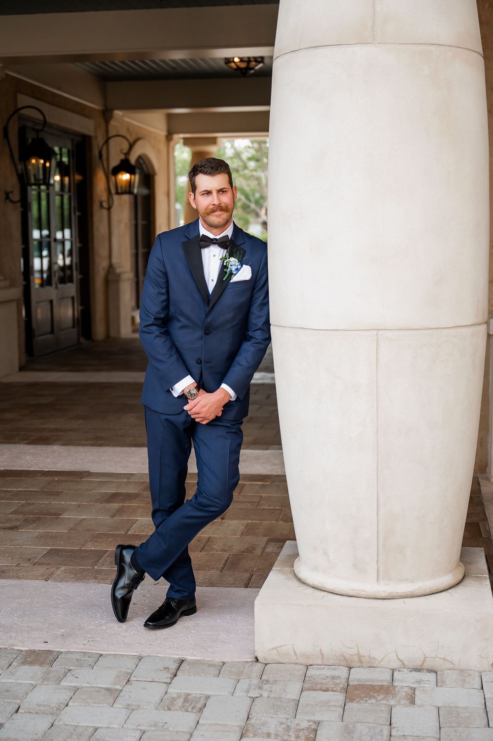 Groom in fitted navy tuxedo poses against white column in elegant venue.
