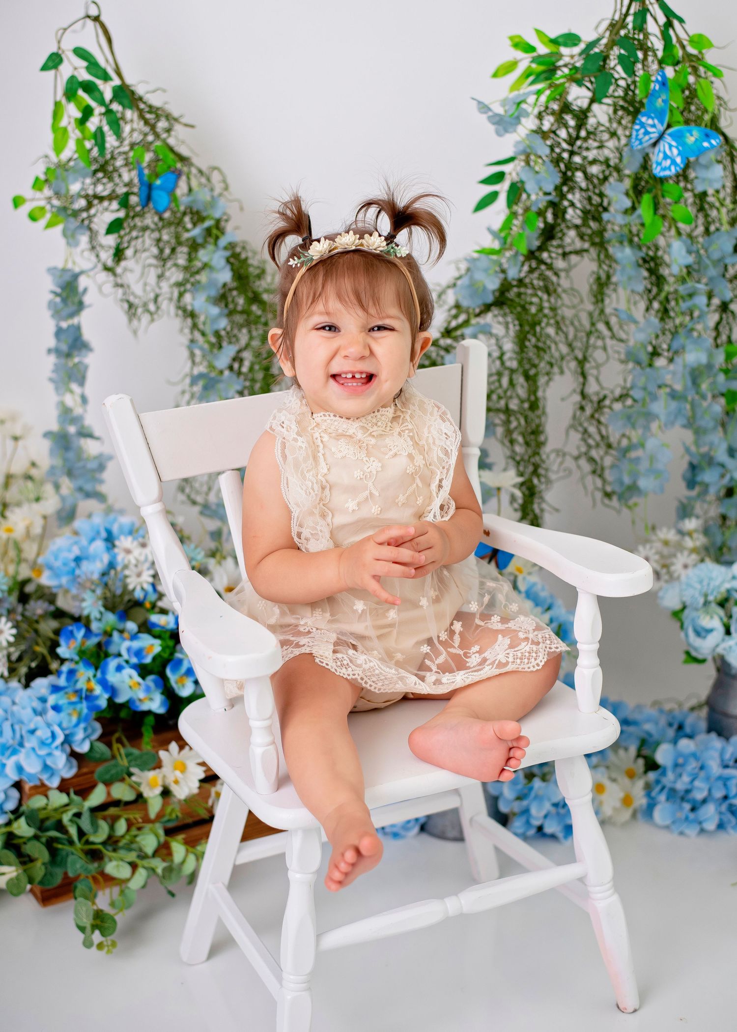 A small child sits in a white wooden chair surrounded by blue flowers and greenery in a photo studio setting.