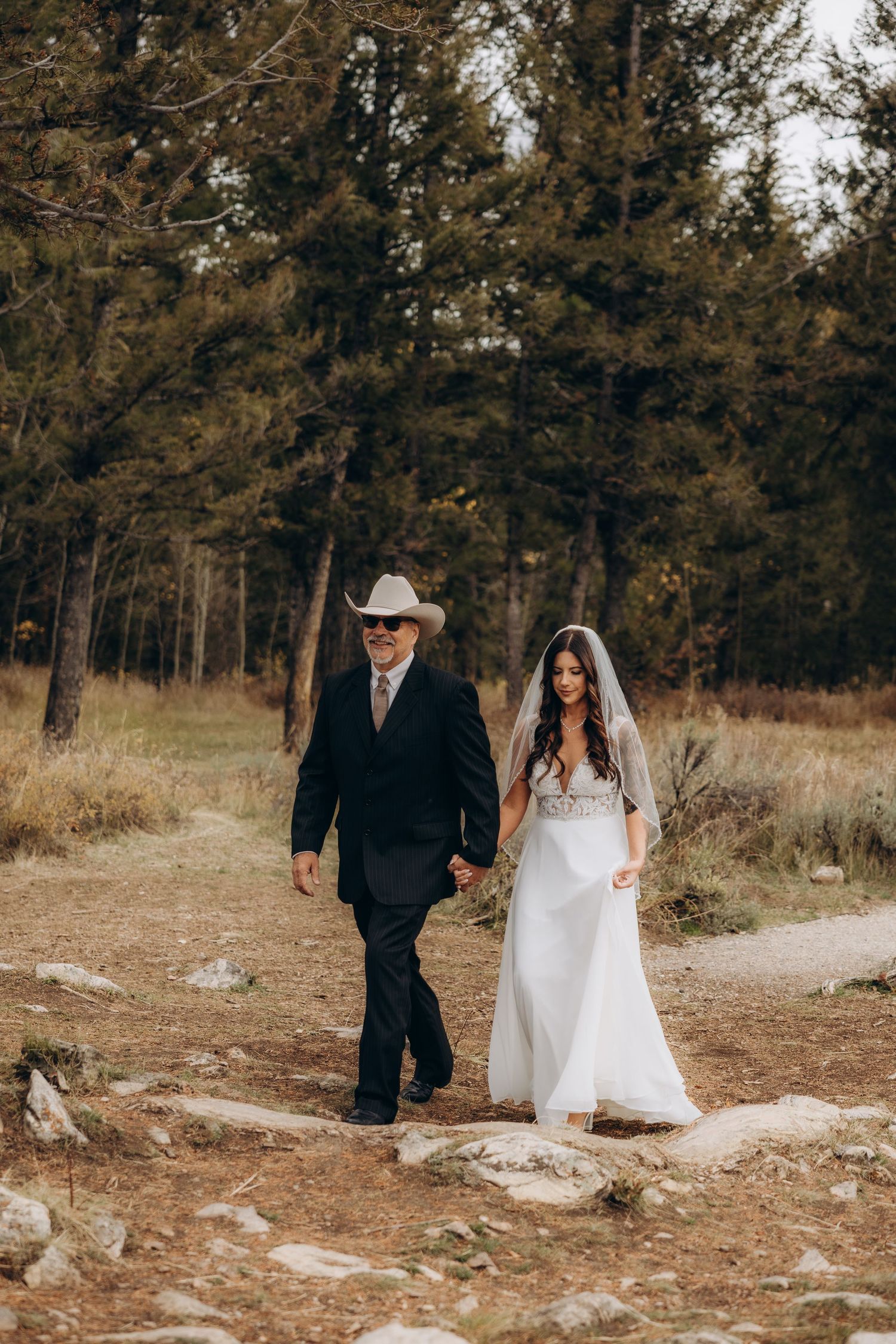 A couple in wedding attire walks through a scenic wooded area with evergreen trees and dry grass in the background.