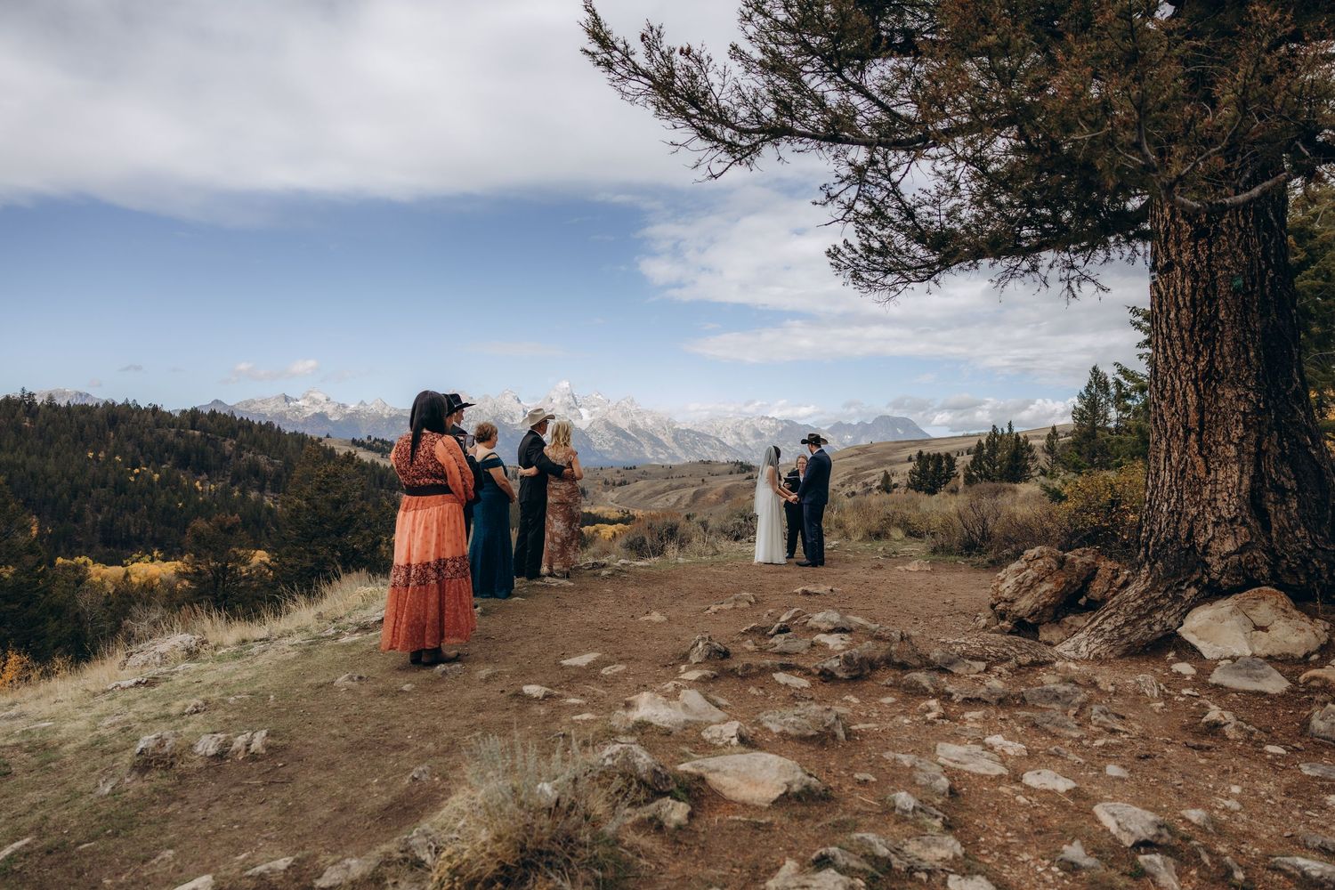 People stand near portable toilets on a scenic mountain overlook with expansive views of rugged peaks and valleys.