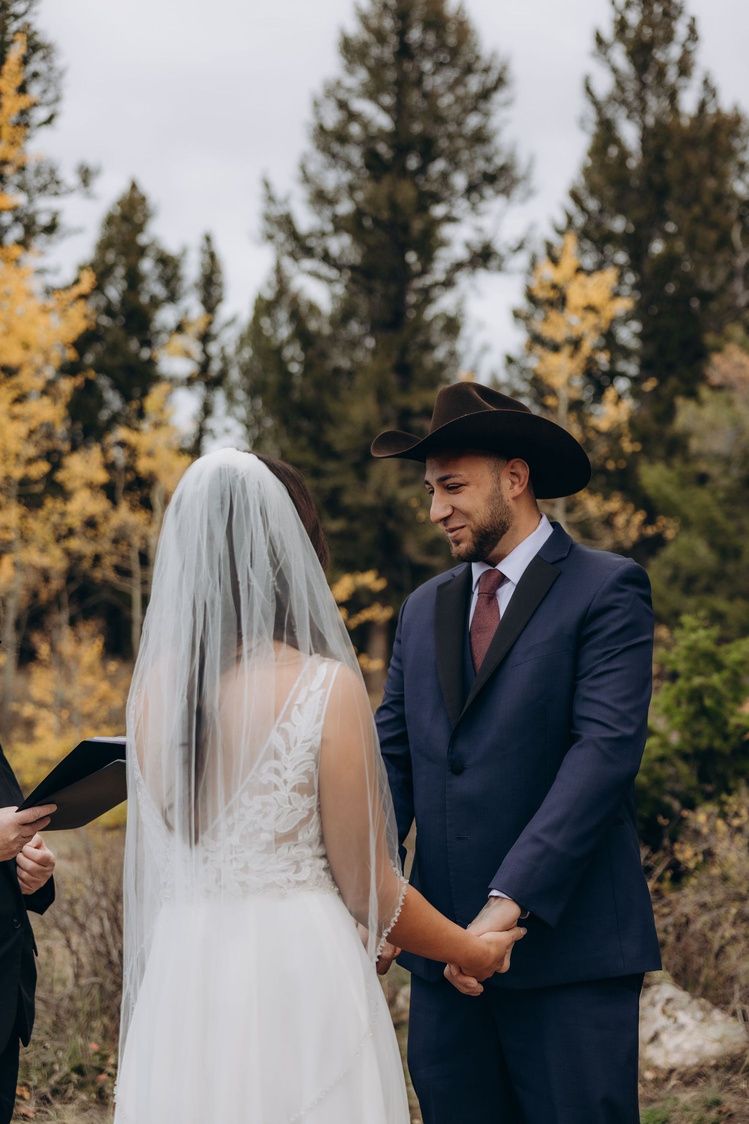A couple exchanges vows during an outdoor wedding ceremony with evergreen trees in the background.