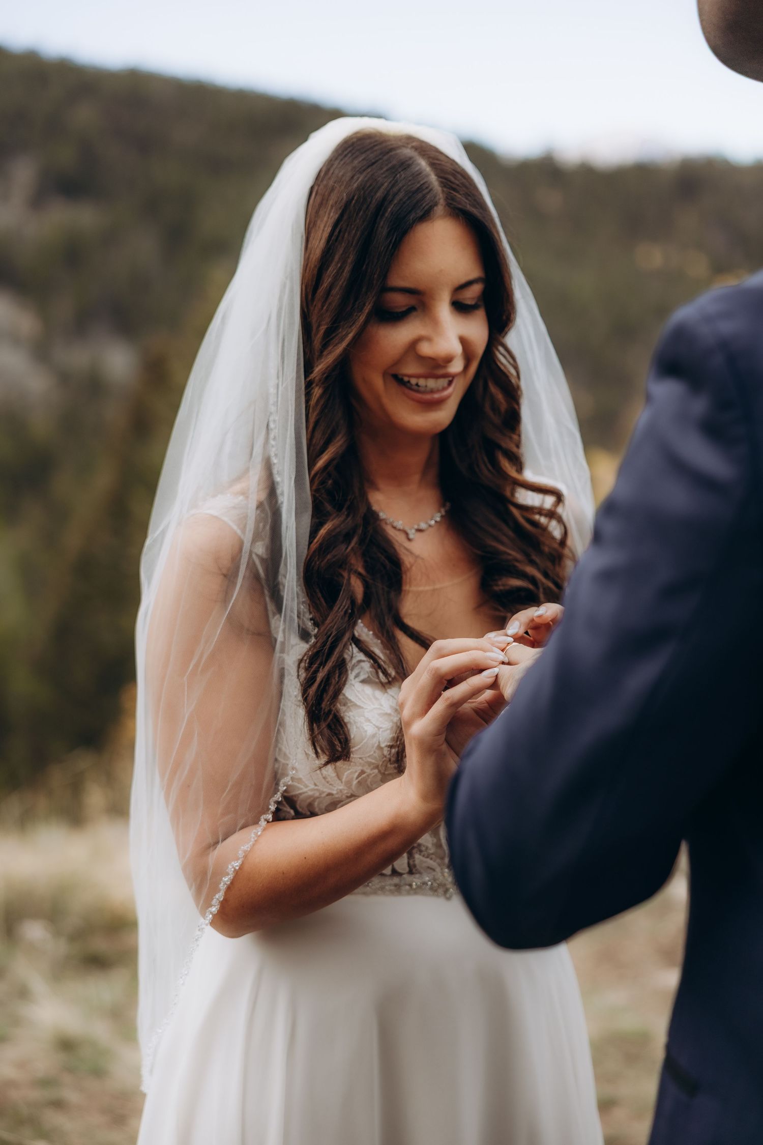 A bride in a white wedding dress and veil smiles while placing a ring during an outdoor ceremony.
