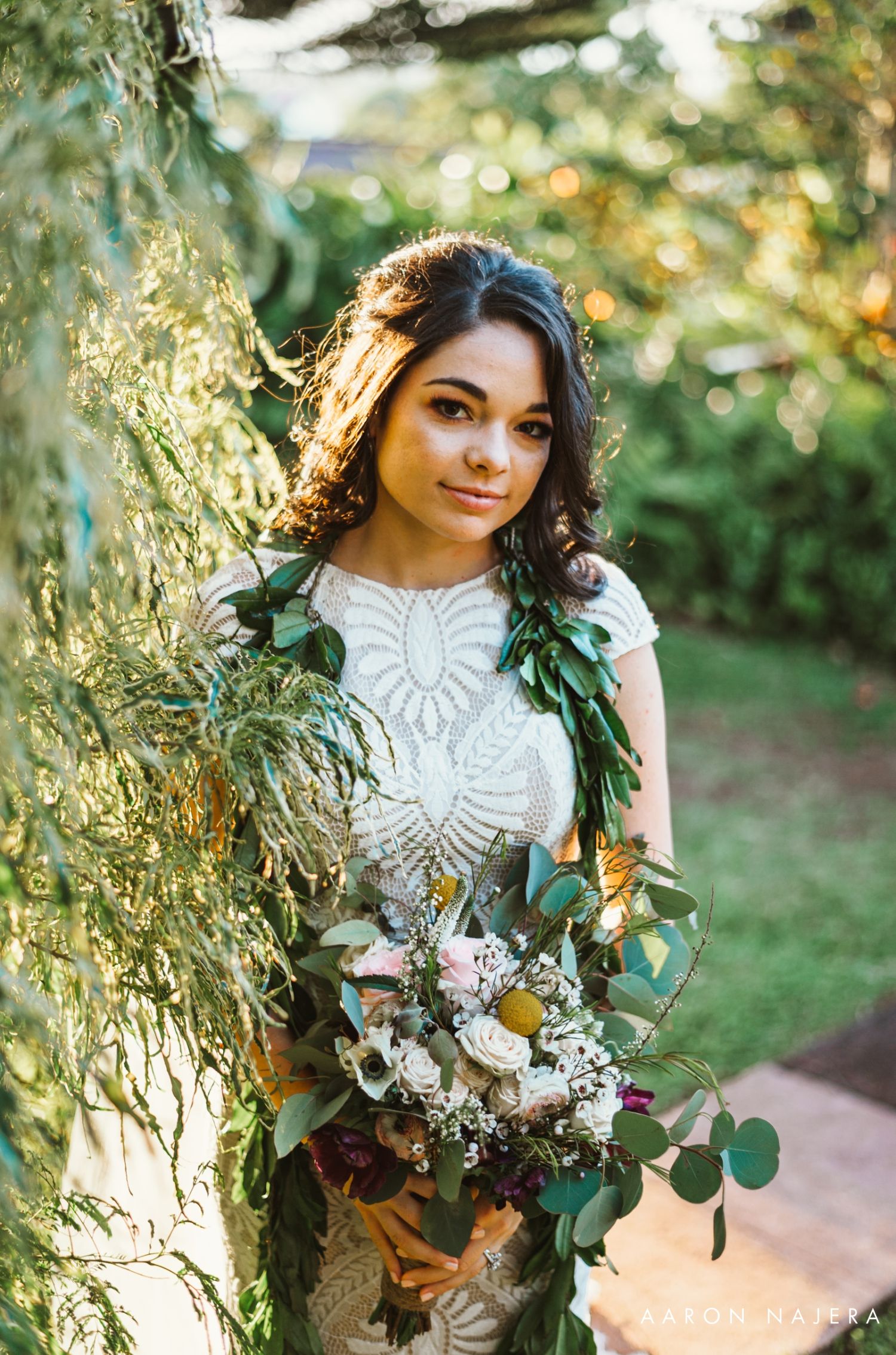 Bride in detailed lace gown poses with natural foliage and bouquet in golden sunlight.