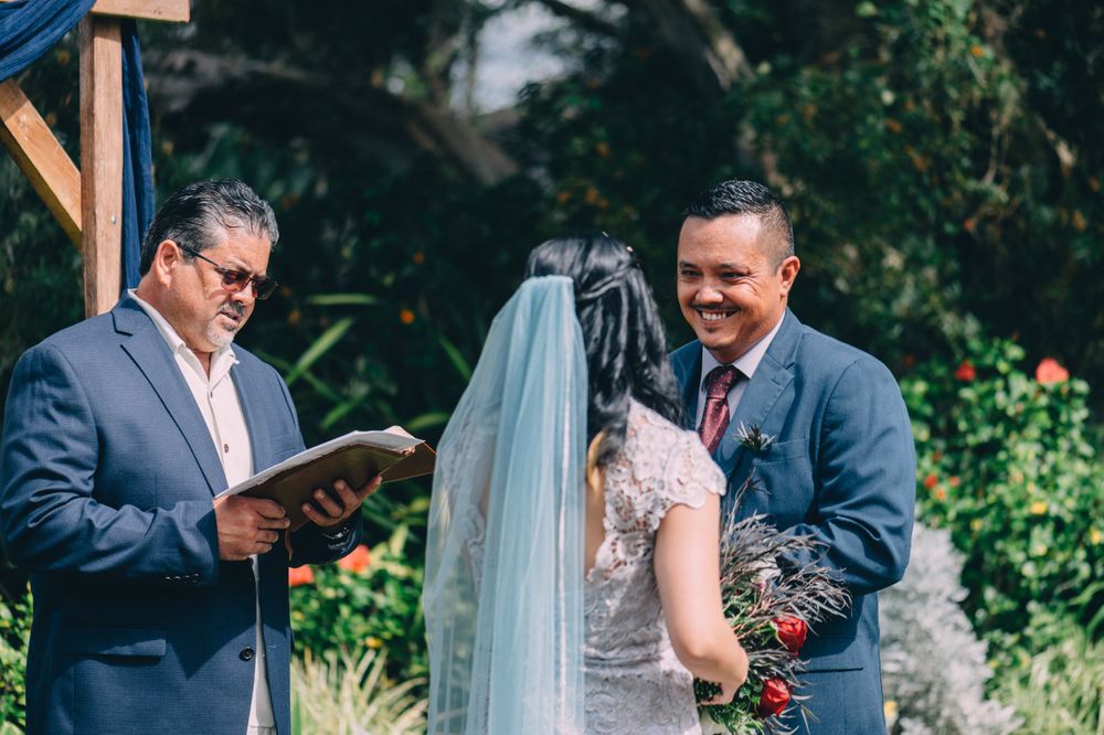 Wedding ceremony under wooden arch with blue draping in garden setting.