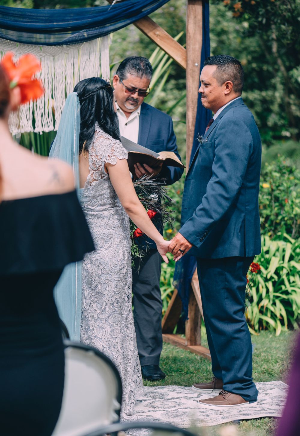 Wedding ceremony under wooden arch with guests seated in garden setting.