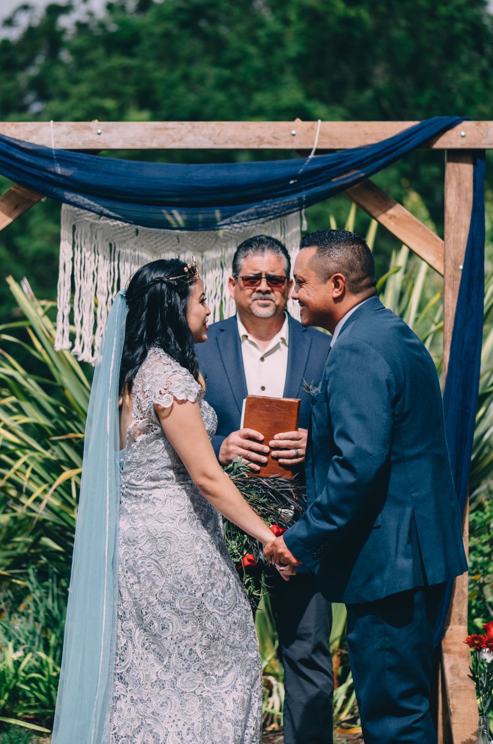 A wedding ceremony takes place under a wooden arch with blue draping and lush greenery in the background.