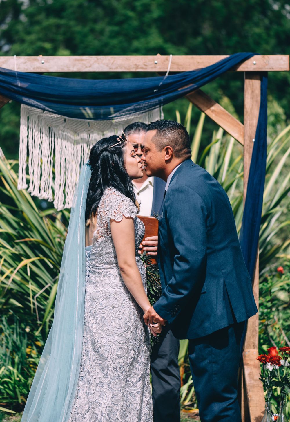 An intimate outdoor wedding ceremony moment captured under a rustic wooden pergola with blue fabric.