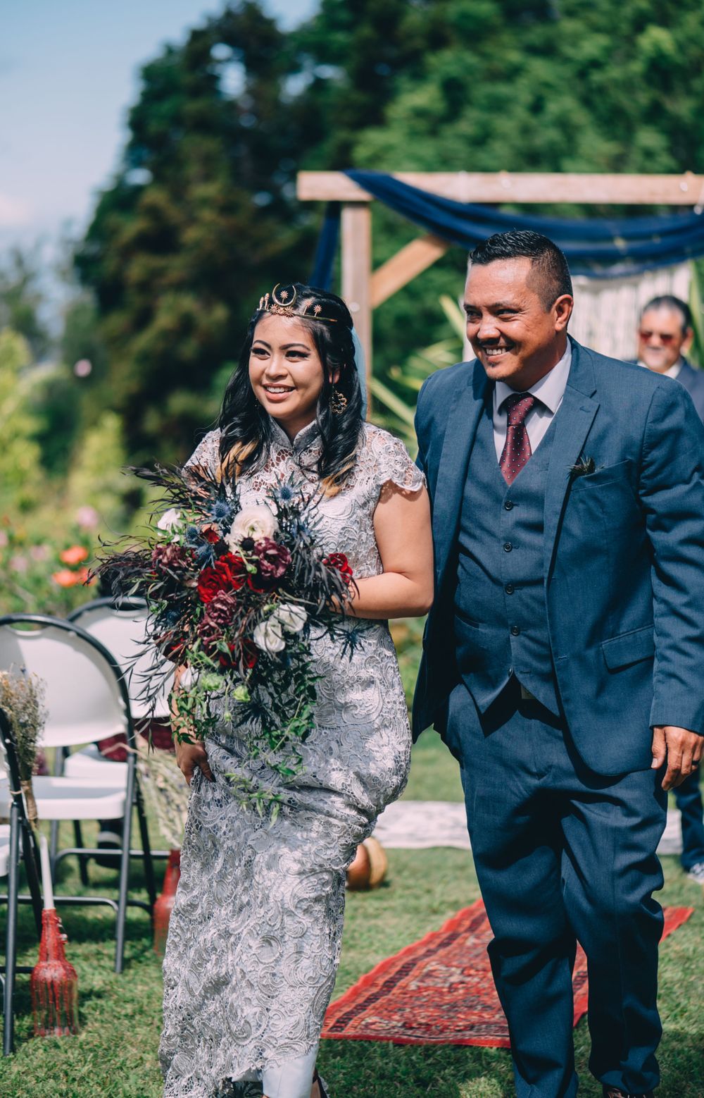 A bride in a silver lace dress holds a dark floral bouquet during an outdoor garden ceremony.