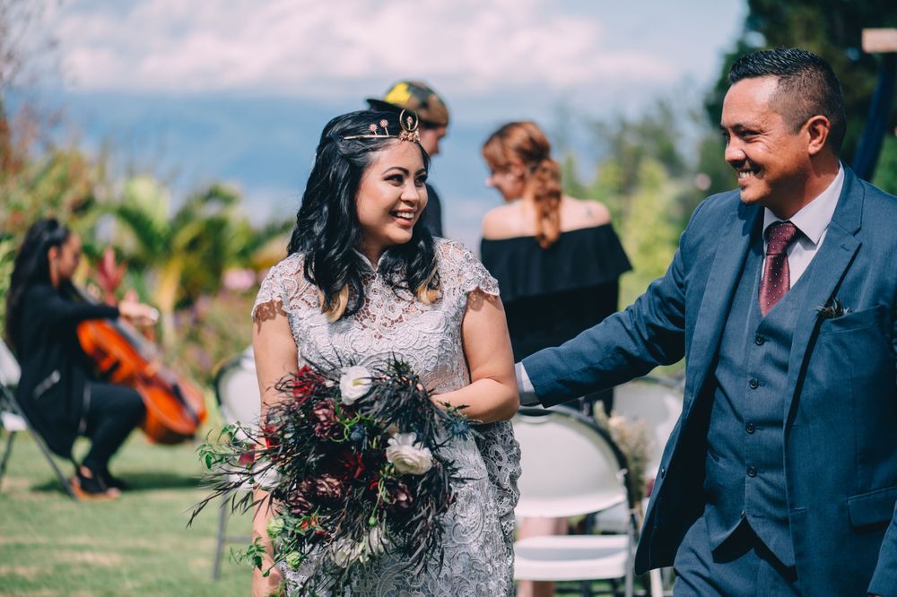 Wedding ceremony participants walk together on a garden path with decorative elements visible.
