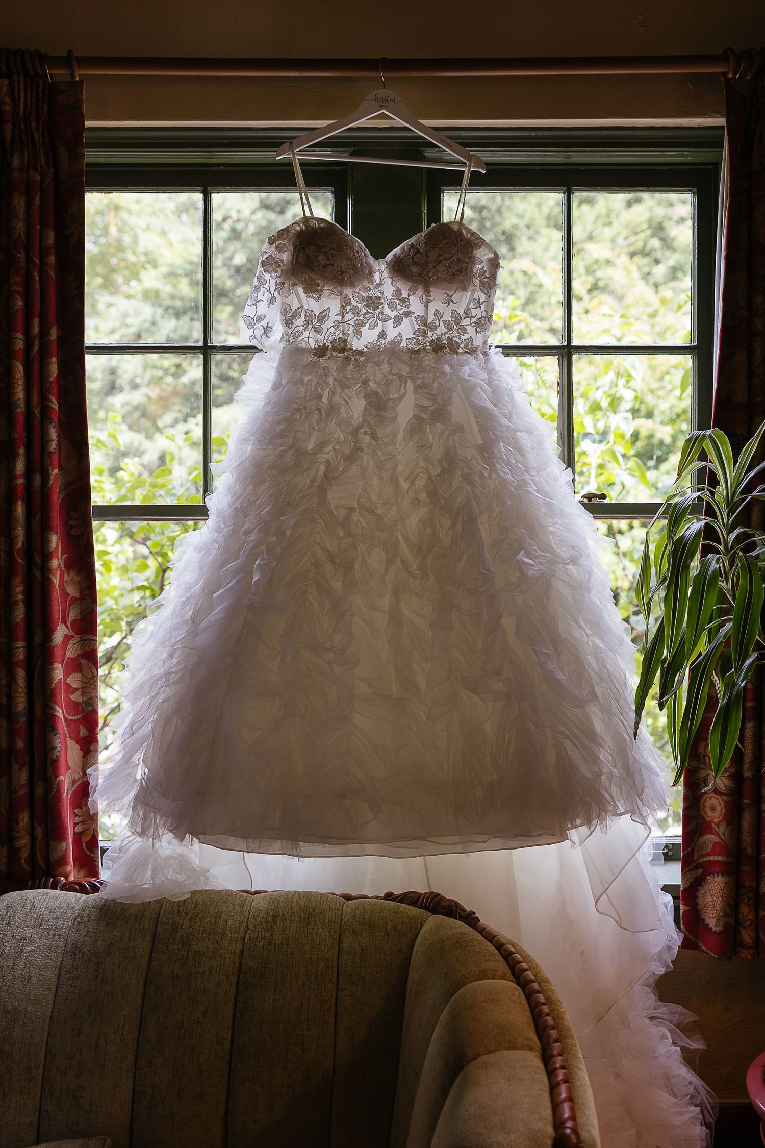 A white wedding dress with lace details hangs in front of windows with natural lighting.