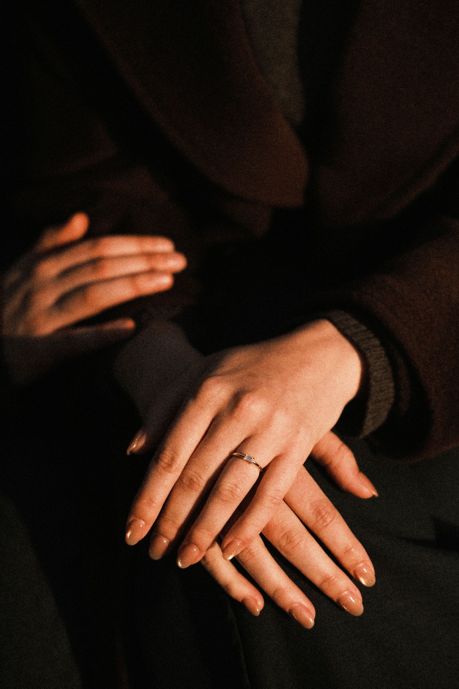 Delicate hands with gold rings rest gently on dark fabric in moody lighting, creating an intimate portrait.