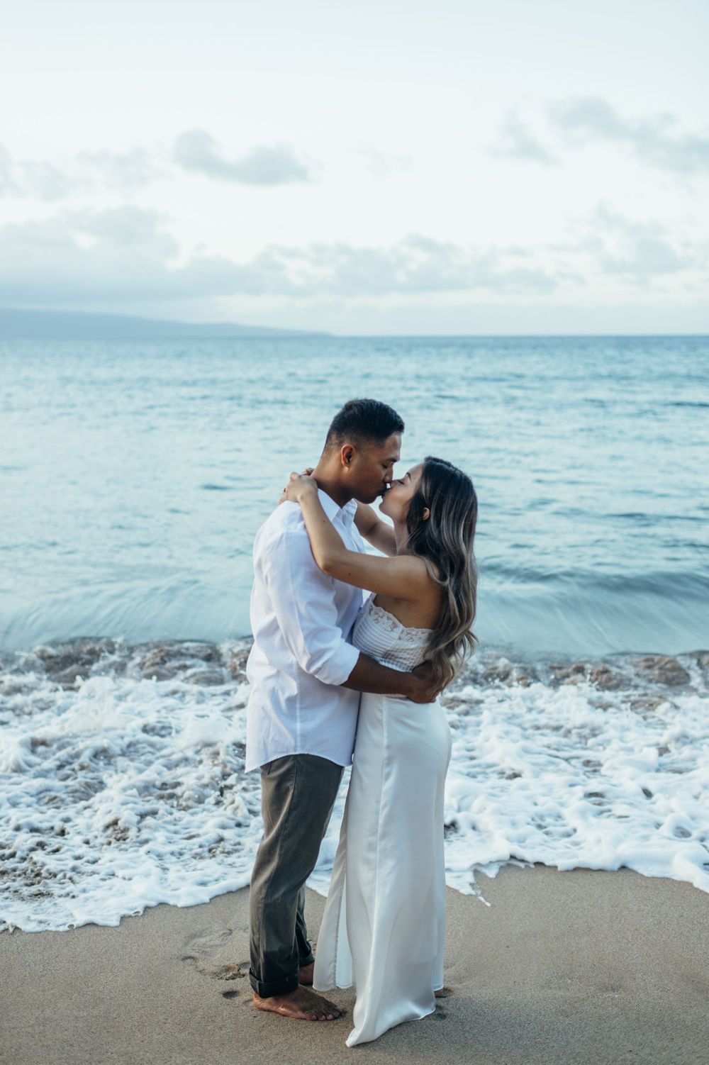 A couple in white embraces on a beach as waves crash behind them.