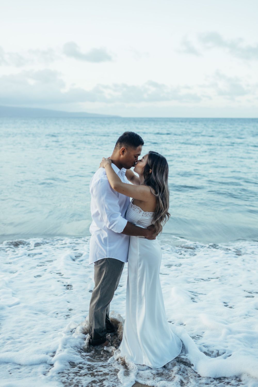 A romantic couple stands in ocean waves wearing white attire.
