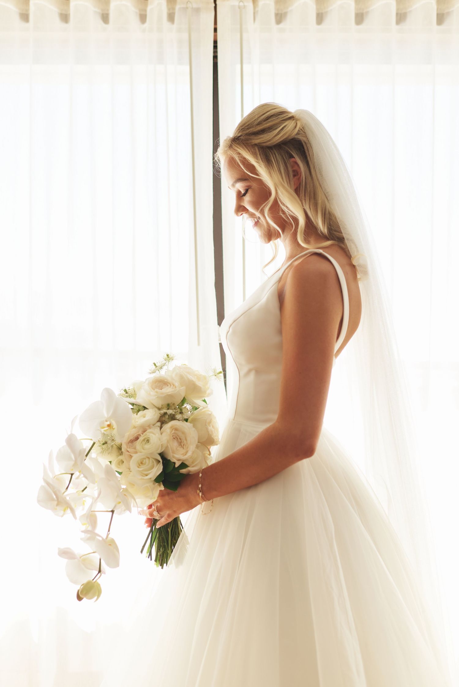 A bride in a white gown poses near sunlit windows with an orchid bouquet.