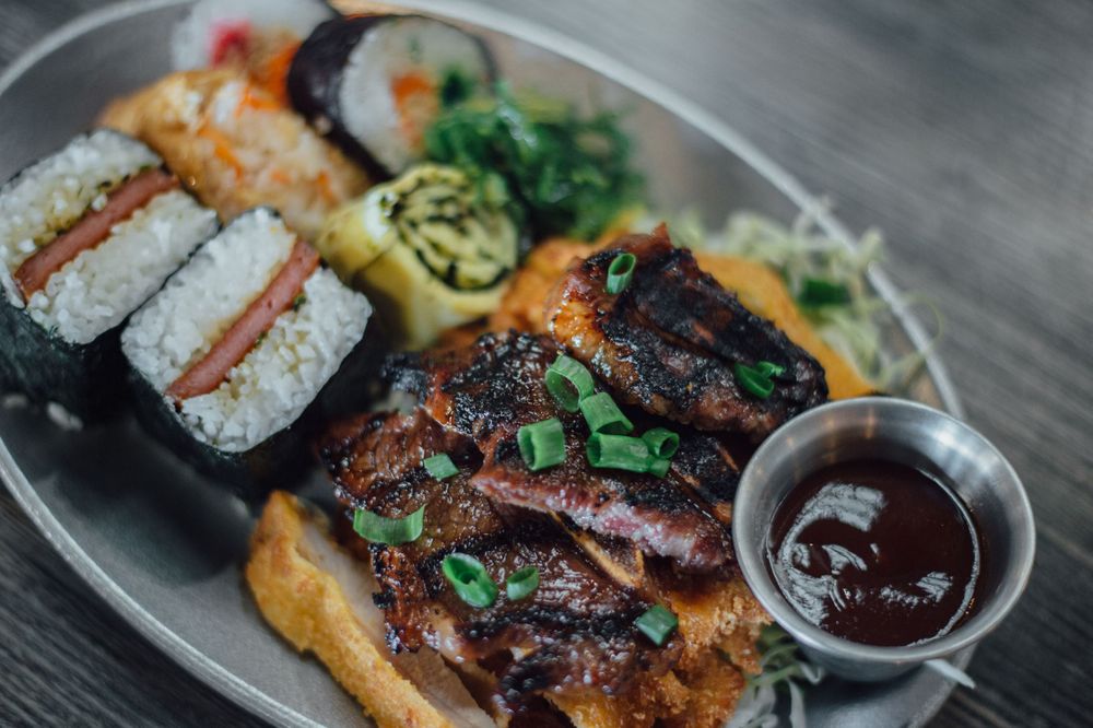 Asian fusion dishes including sushi rolls and grilled meats with dipping sauce on white plates.