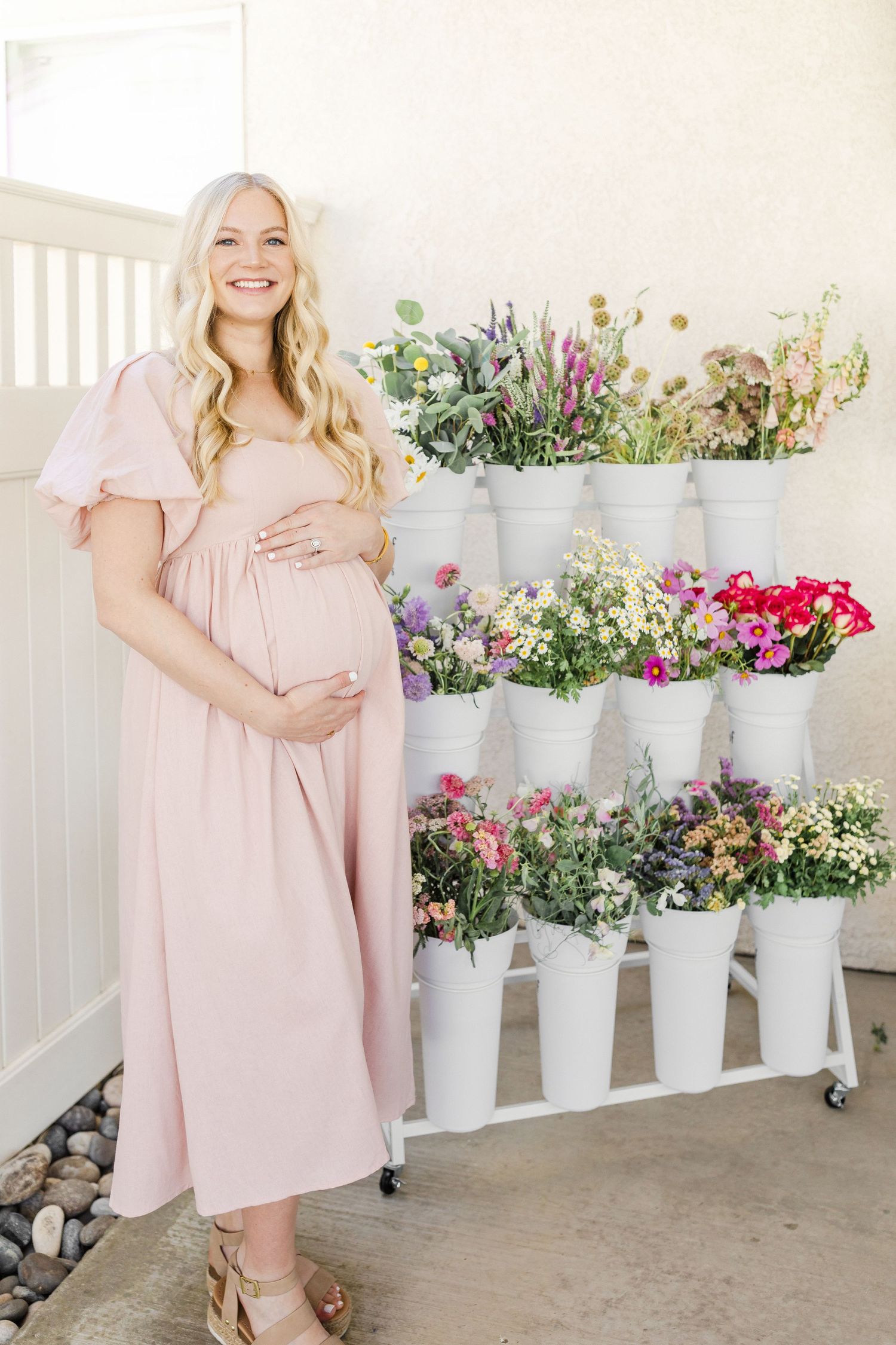 Mom to be in a blush pink dress poses in front of white planters filled with colorful spring flowers and greenery captured by Chelsee Rawe