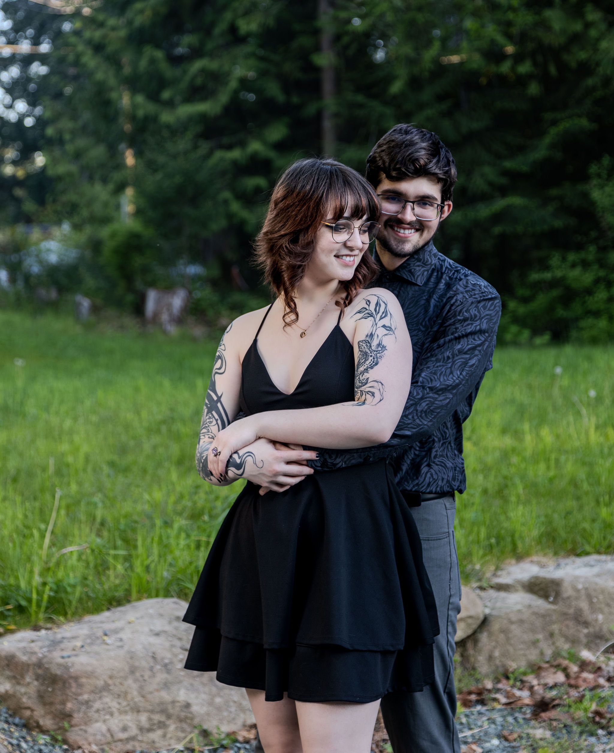 Tattooed couple posing together outdoors, woman in black dress, man in dark patterned shirt.