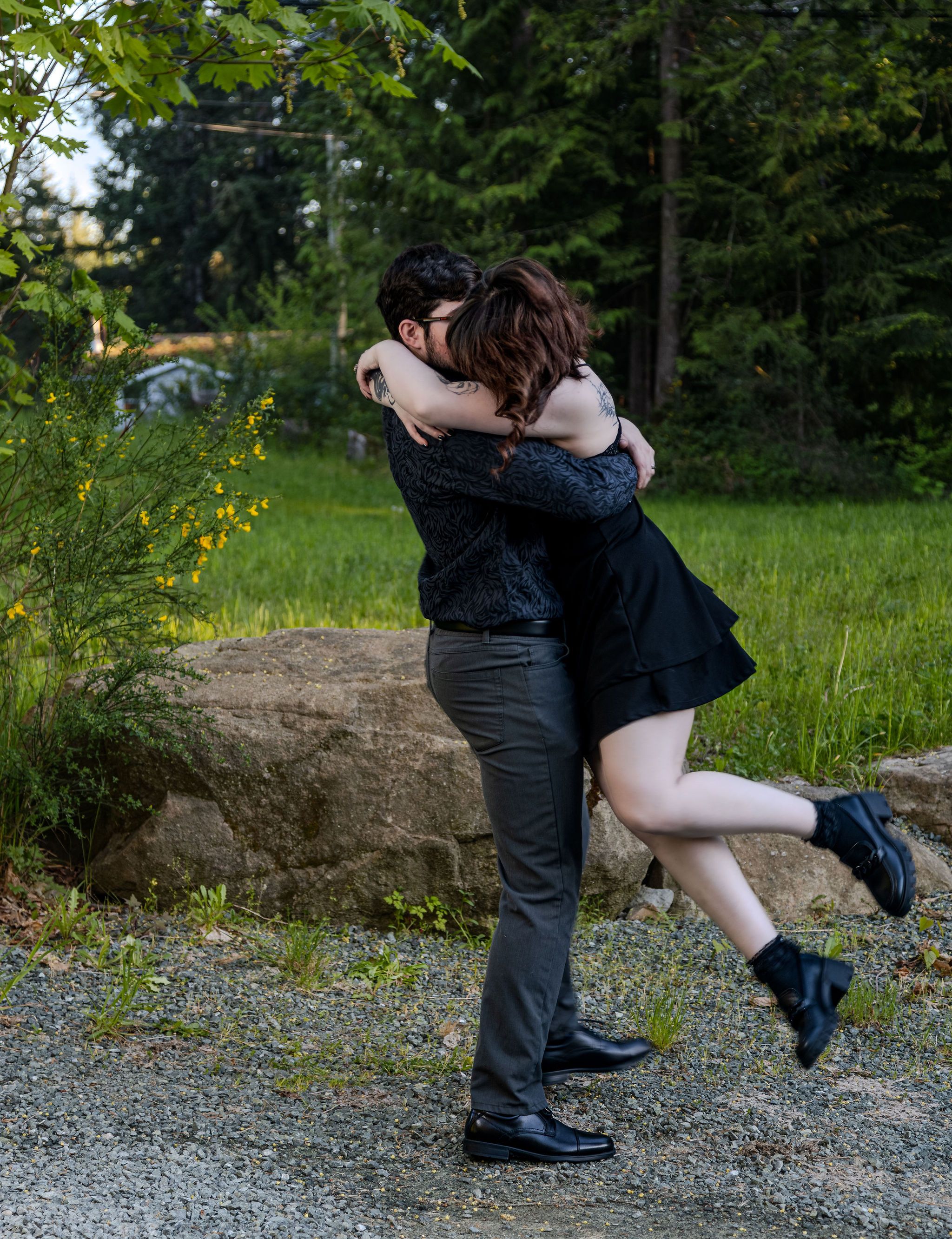 Man joyfully lifts woman in black dress during outdoor couples photo session near forest.